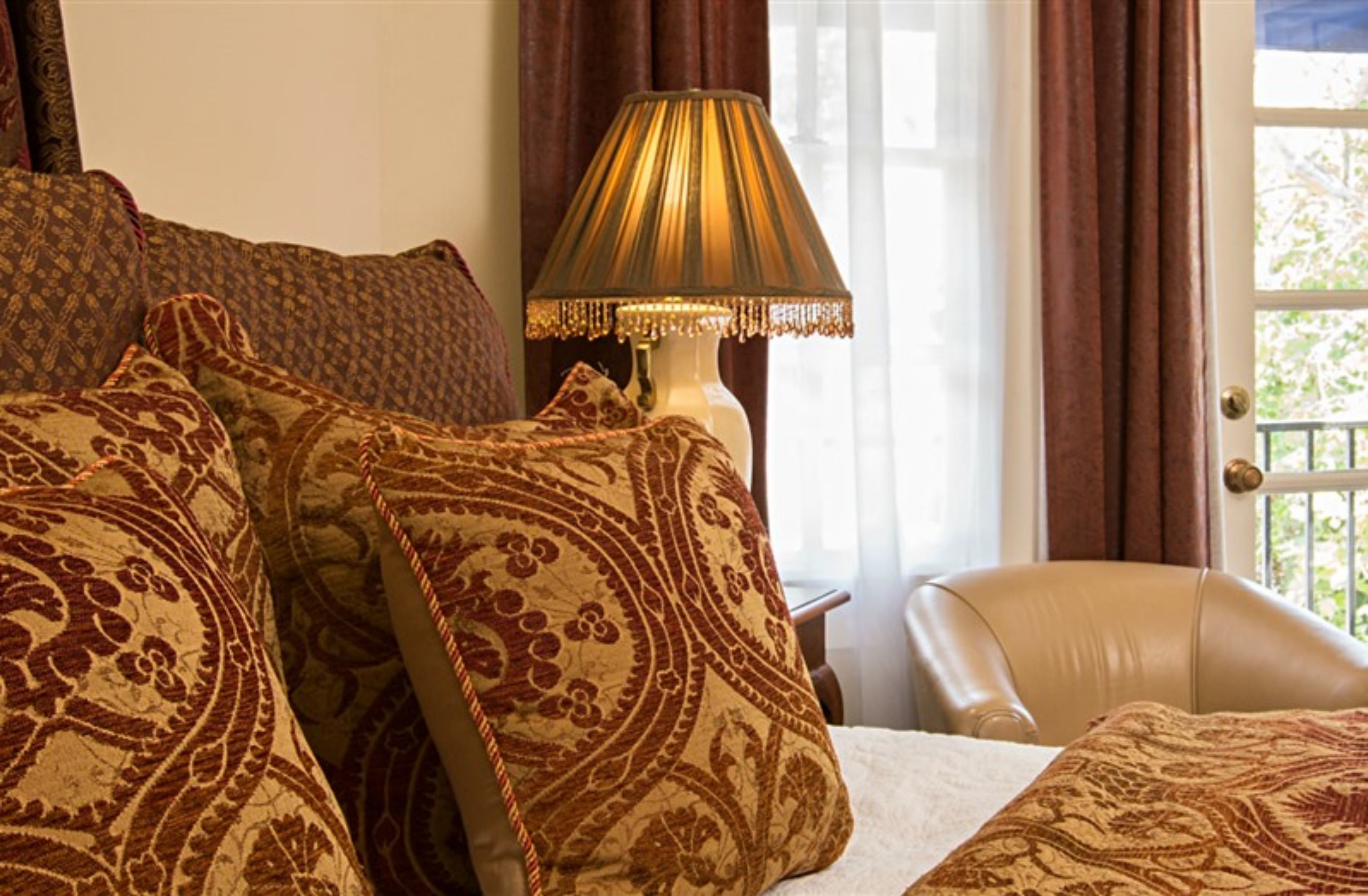 A bedroom with a bed, patterned pillows, a lamp, a chair, and a view through a window with curtains.