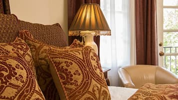 Un dormitorio con una cama, almohadones estampados, una lámpara, una silla y vistas a través de una ventana con cortinas.