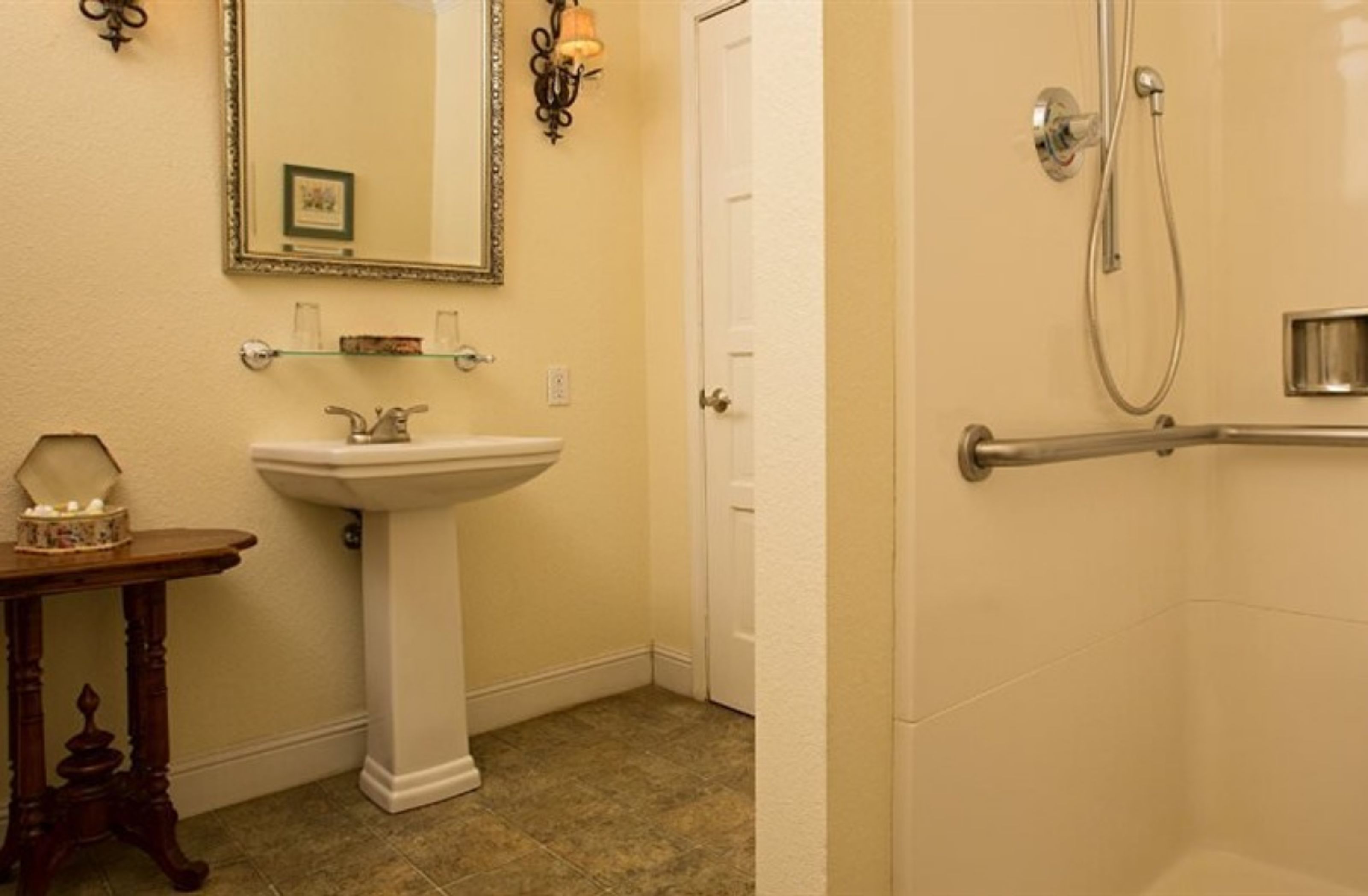 A bathroom with a pedestal sink, a mirror, a shower with a grab bar, and a door.