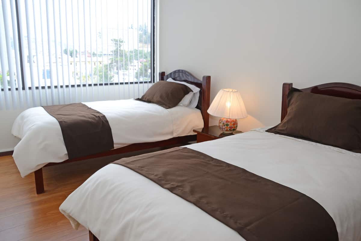 Dos camas individuales con cabeceros de madera, ropa de cama blanca y marrón, una lámpara de noche y una ventana con persianas.
