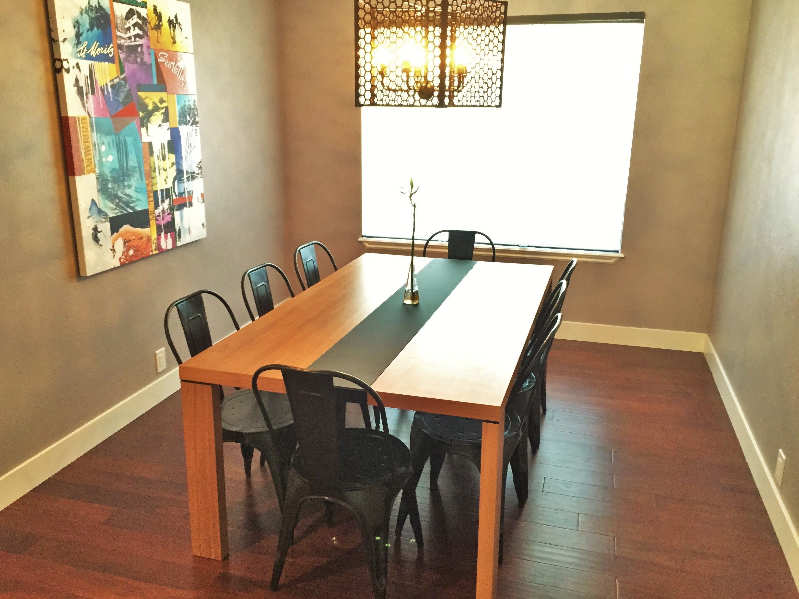 Een eethoek met een lange houten tafel, zes zwarte stoelen, een kroonluchter en een groot raam.