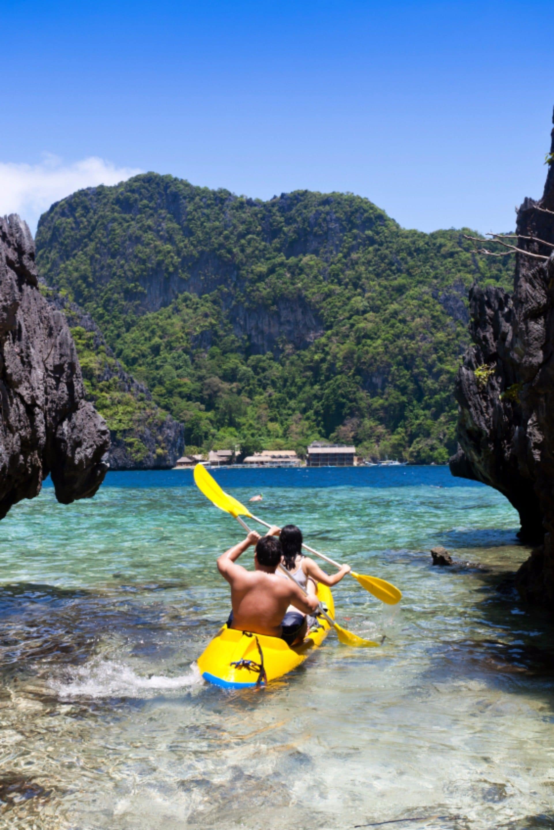 A kayak with a person paddling through clear water between rocky formations, with a forested mountain in the background.