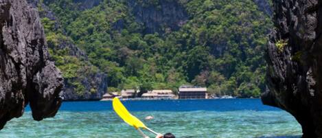 A kayak with a person paddling through clear water between rocky formations, with a forested mountain in the background.