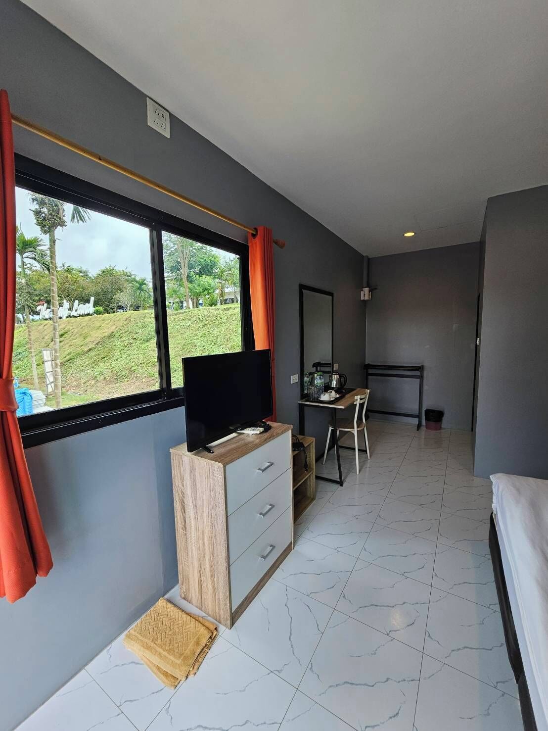 A modern hotel room with a large window, a TV on a wooden cabinet, a desk with a chair, and a bed.