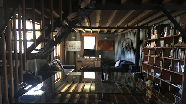 A rustic interior with a glass table, leather sofas, a bookshelf, and a staircase.