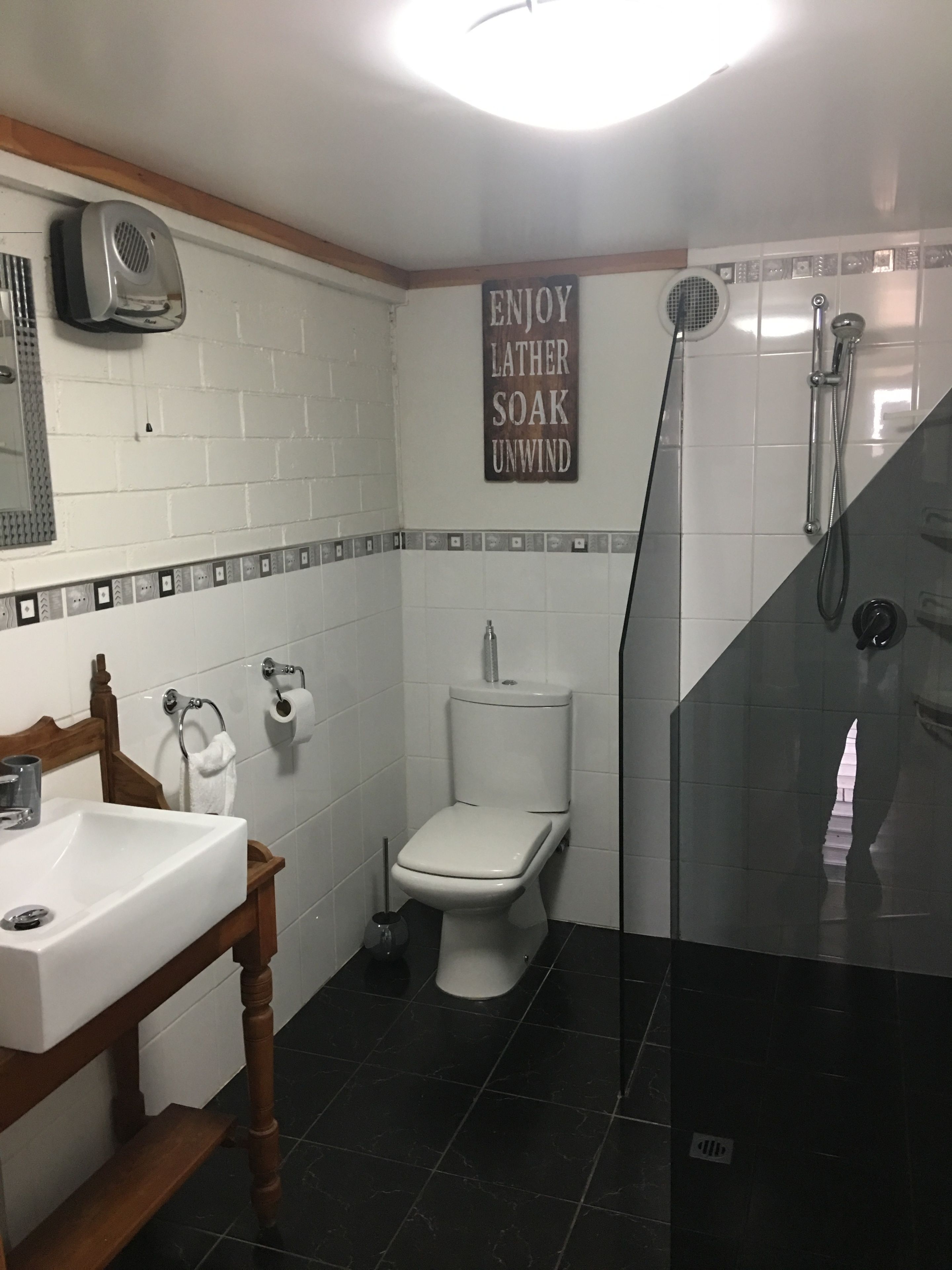 A bathroom with a white sink, toilet, and shower. There is a sign on the wall that says "ENJOY LATHER SOAK UNWIND".