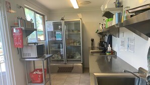 Eine Küche mit Edelstahlgeräten, inklusive Kühlschrank und Spüle. Regale mit verschiedenen Gegenständen sowie ein Feuerlöscher an der Wand.