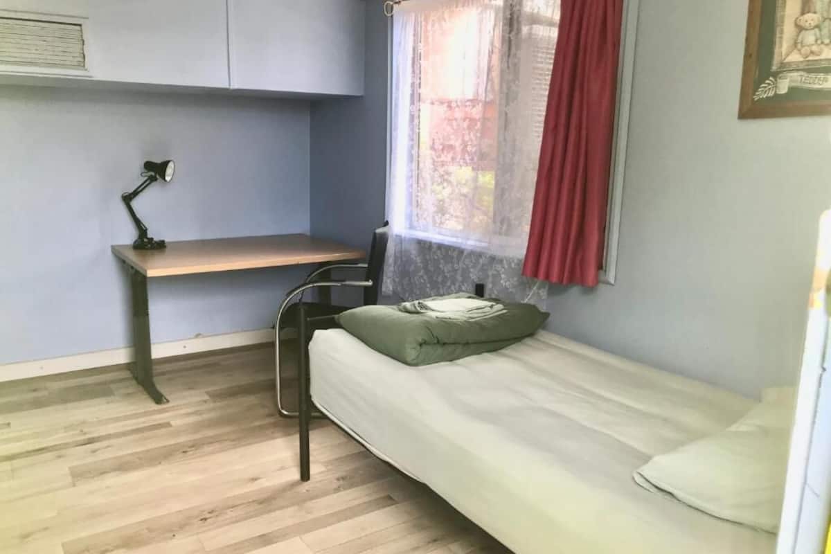 Quarto pequeno com uma cama de solteiro, escrivaninha e cadeira, janela com cortina vermelha e piso de madeira.