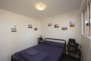 Un lit simple avec un matelas et des oreillers bleus, une petite table de chevet avec une lampe et une fenêtre avec des stores.
