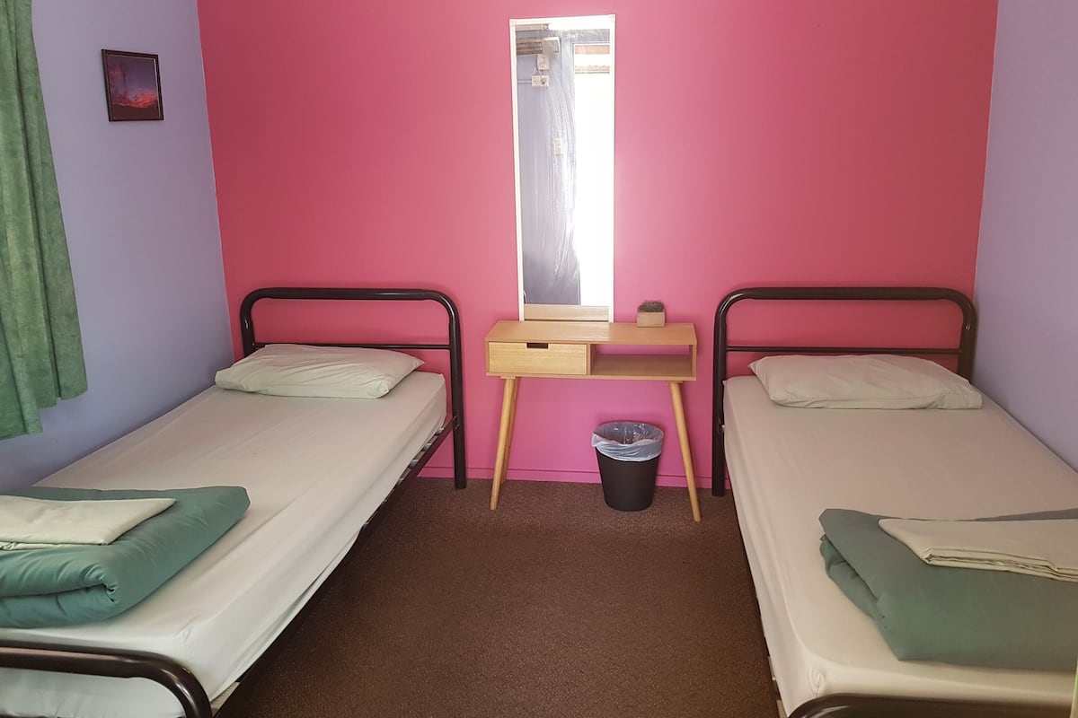 Quarto com duas camas de solteiro, uma escrivaninha, um espelho e uma janela.