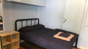 Ein kleines Zimmer mit einem Einzelbett, Holzboden, einem Metallbettgestell, einer dunkel gefärbten Bettdecke und einem beigen Kopfkissen. Eine weiße Tür und ein kleines Holzregal sind links zu sehen.