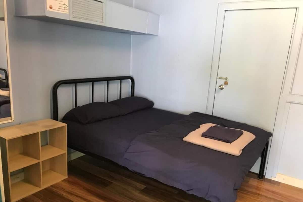 Quarto pequeno com uma cama de solteiro, chão de madeira, estrutura de cama de metal, colcha escura e travesseiro bege. Há uma porta branca e um pequeno móvel de madeira à esquerda.