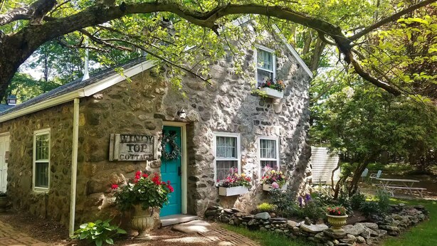 Une petite maison en pierre avec une porte turquoise, une enseigne indiquant « Windy Top Cottage », et des jardinières aux fenêtres.