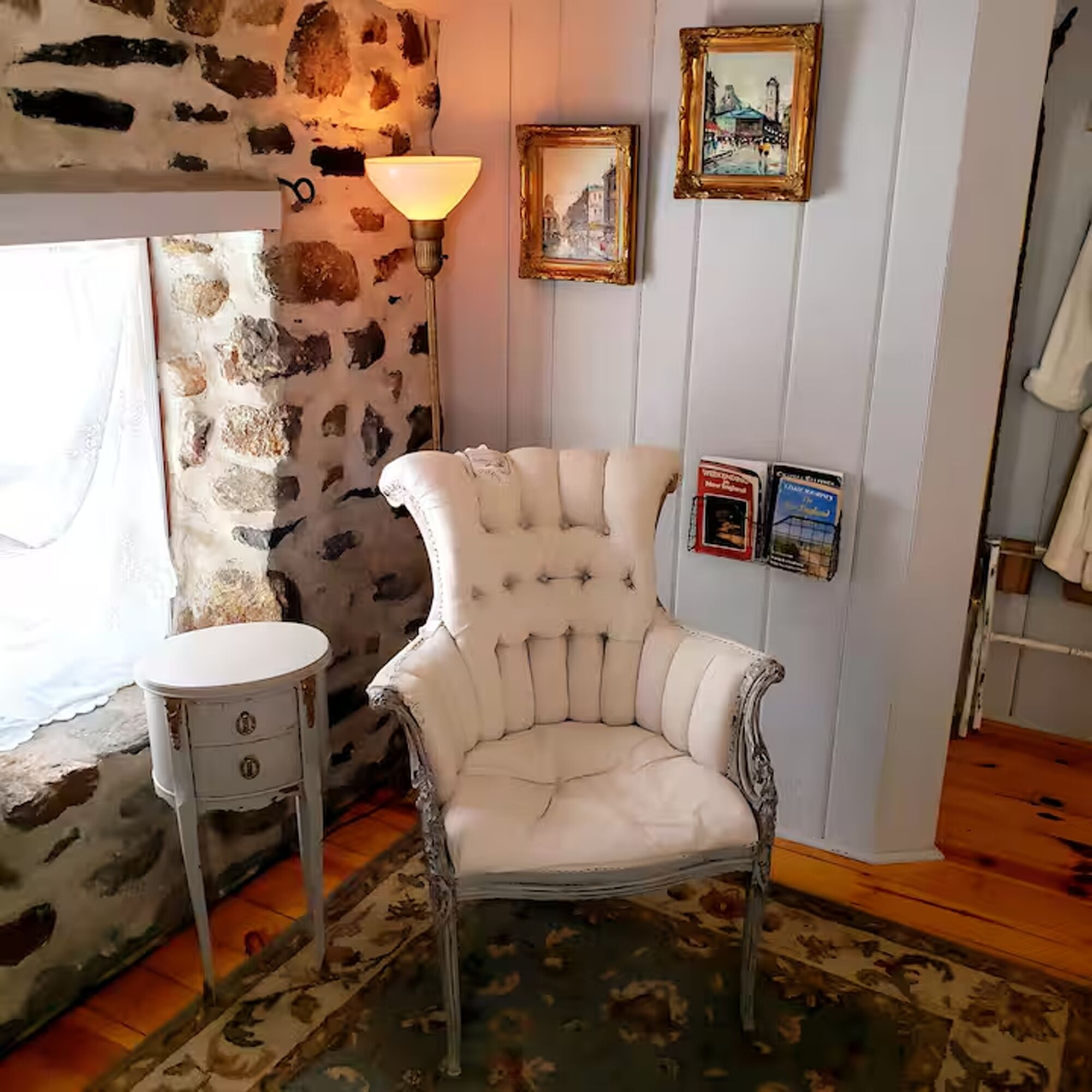 A room with a stone wall, a white armchair, a small round table, and framed pictures on the wall.