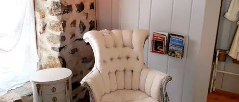A room with a stone wall, a white armchair, a small round table, and framed pictures on the wall.