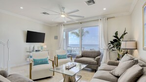 Un salon spacieux avec une table basse en verre, des sièges confortables, un ventilateur de plafond et une grande fenêtre donnant sur l’océan.