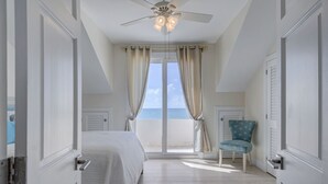 Une chambre à coucher avec un lit, une chaise, un ventilateur de plafond et une vue sur la mer grâce à de grandes fenêtres.