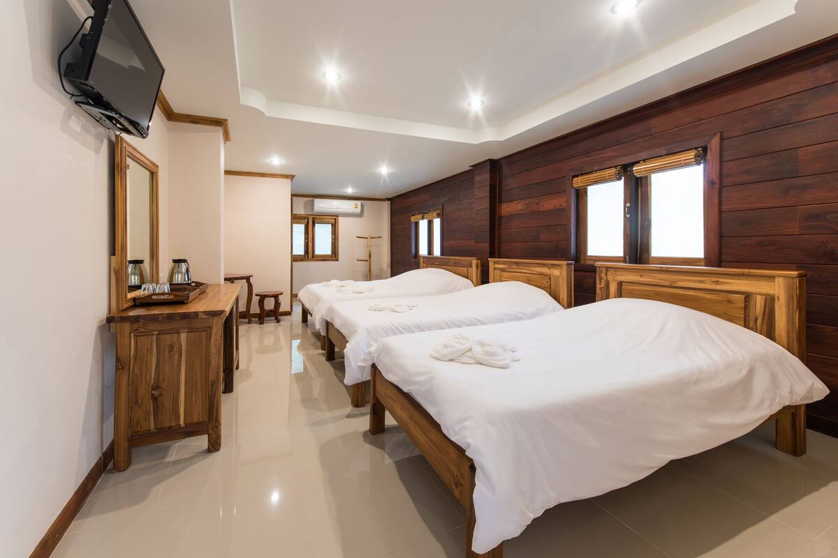 A hotel room with three wooden beds, a wooden dresser, a television mounted on the wall, and a window with blinds.