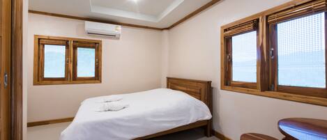 A bedroom with a bed, wooden headboard, two windows with blinds, a wooden bench, and an air conditioning unit.