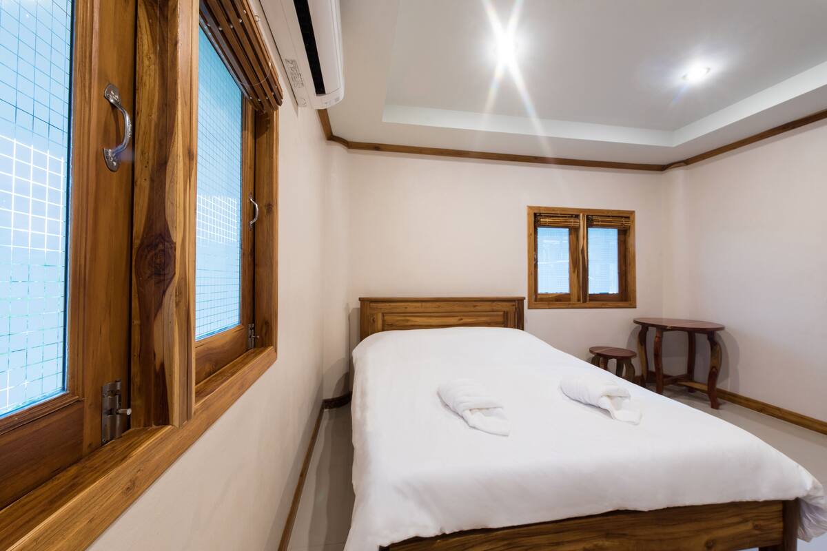 A hotel room with a wooden bed, a small wooden table, a window with blinds, and an air conditioning unit.