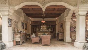 An ornate lobby with a high ceiling, intricate columns, and a central fireplace. There are wicker chairs and a small table in the foreground.