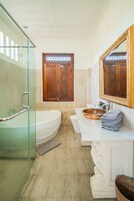 A modern bathroom with a glass shower enclosure, a freestanding bathtub, a large mirror, and a wooden door.