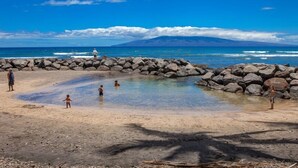 Ein Strand mit einer Felsbucht, klarem, blauem Wasser und einem Berg im Hintergrund.