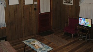 Una habitación con paredes de paneles de madera, una puerta con cortina blanca, un televisor sobre un mueble de madera, una silla roja y una mesita con una revista.