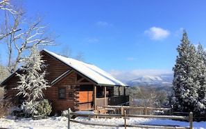 Une cabane en bois dont le toit est recouvert de neige, entourée d’arbres et de montagnes également enneigés, qui se dressent à l’arrière-plan.