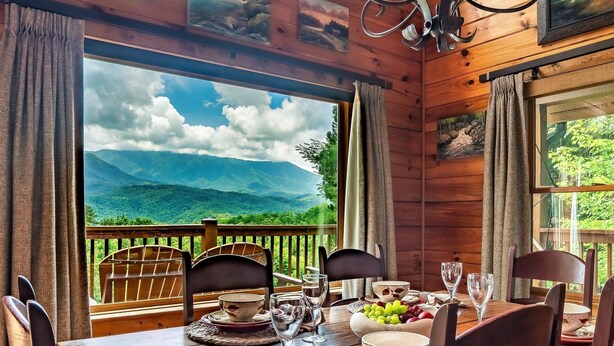 Ein Holztisch mit Stühlen, eine Obstschale und ein Weinglas auf einem Balkon mit Blick auf eine gebirgige Landschaft.