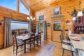 A wooden cabin interior with a dining area, kitchen, and a large window offering a view of the mountains.