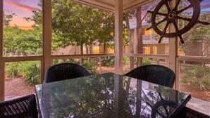A covered outdoor seating area with a glass table and chairs, a ship's wheel decoration, and a view of trees and buildings.