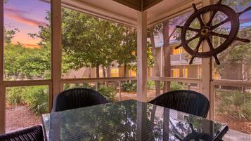 A covered outdoor seating area with a glass table and chairs, a ship's wheel decoration, and a view of trees and buildings.