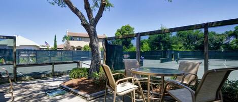 An outdoor patio with wicker chairs and a glass table, surrounded by a wooden fence and a tree.