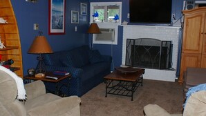 Ein Wohnzimmer mit einem blauen Sofa, zwei Sesseln, einem Couchtisch, einem Kamin, einem Fernseher und einem Fenster mit Jalousien.