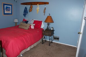 A bedroom with a red bedspread, a wooden headboard, a nightstand with a lamp, and a wall-mounted surfboard.