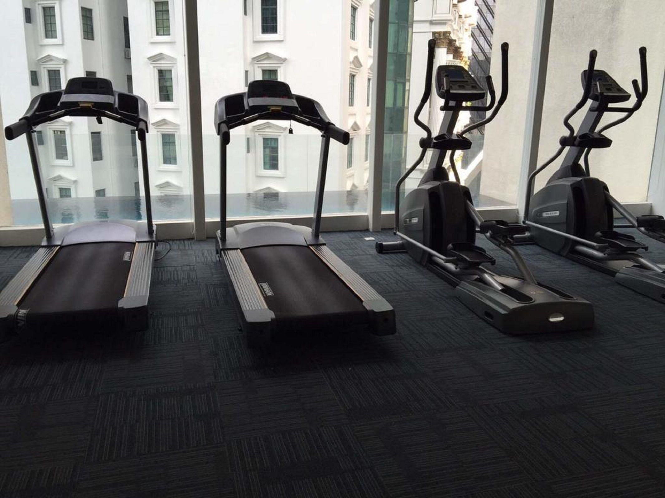 Une salle de sport équipée de tapis de course et d’appareils elliptiques, avec de grandes fen êtres donnant sur des immeubles.