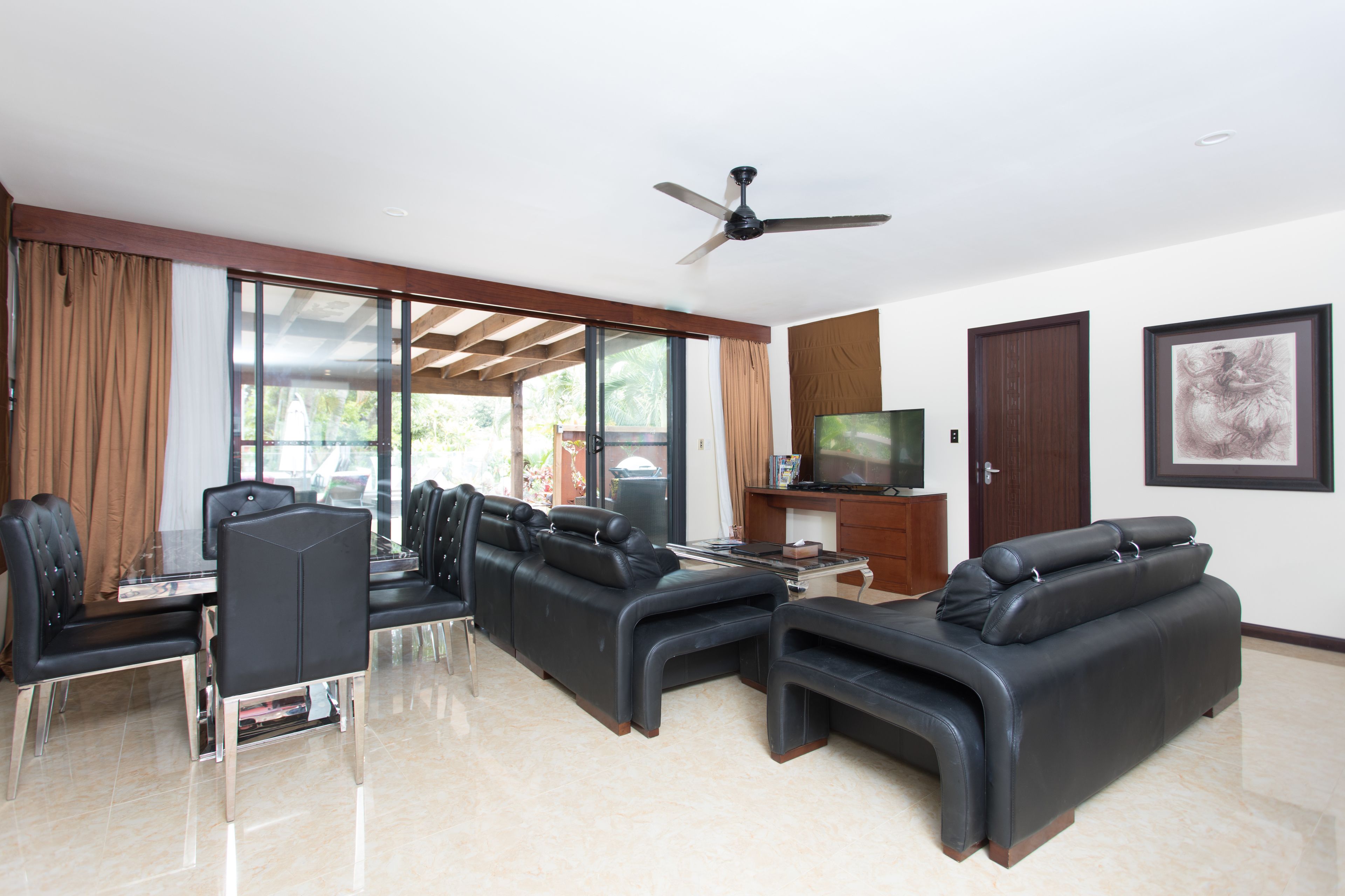 A modern living room with a glass dining table, black leather sofas, a ceiling fan, and a TV on a wooden cabinet.