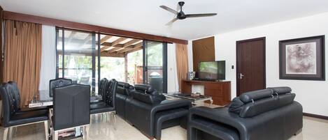 A modern living room with a glass dining table, black leather sofas, a ceiling fan, and a TV on a wooden cabinet.