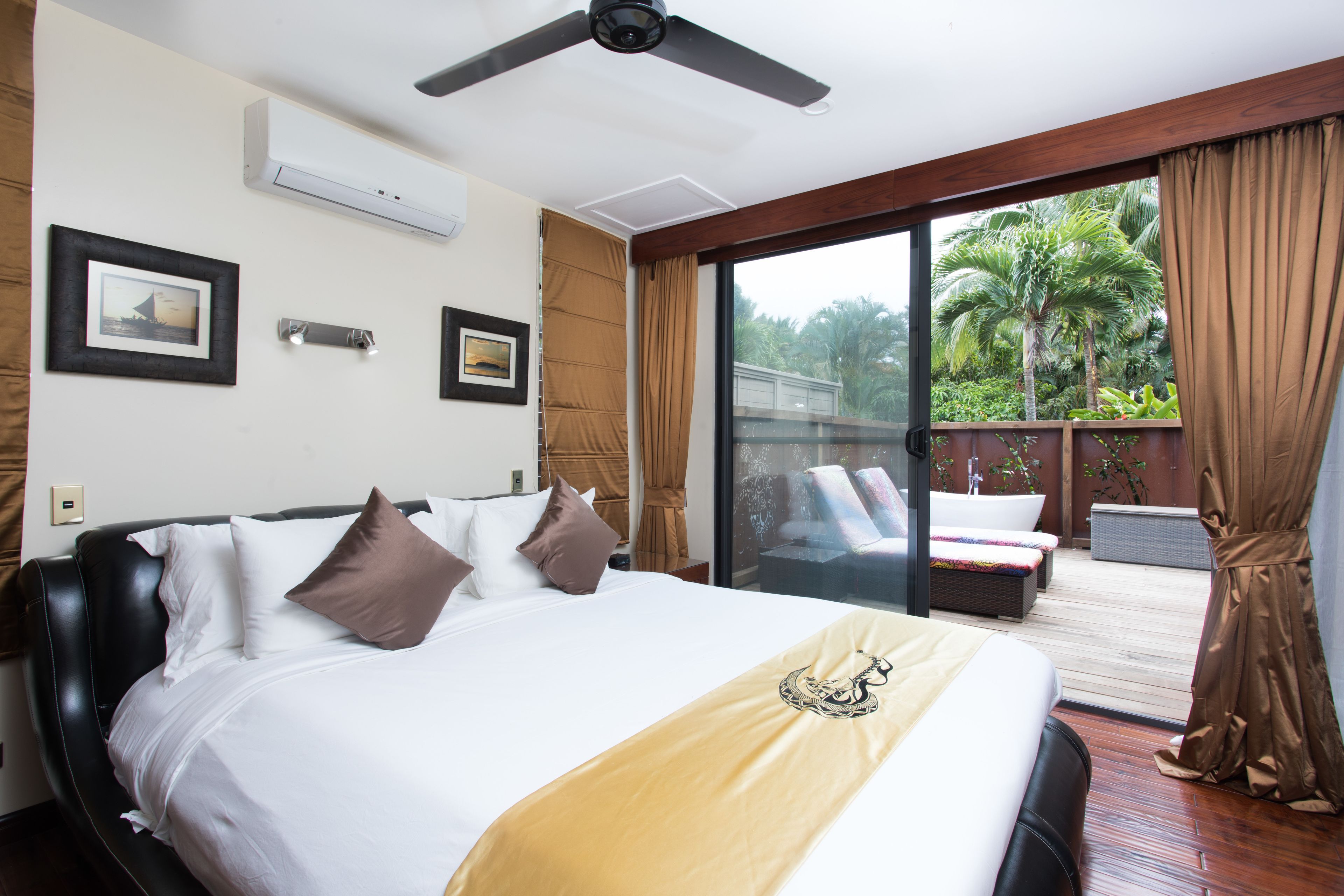 Quarto de hotel com uma cama grande, um ventilador de teto e uma porta de vidro deslizante que dá para uma varanda com móveis de exterior.