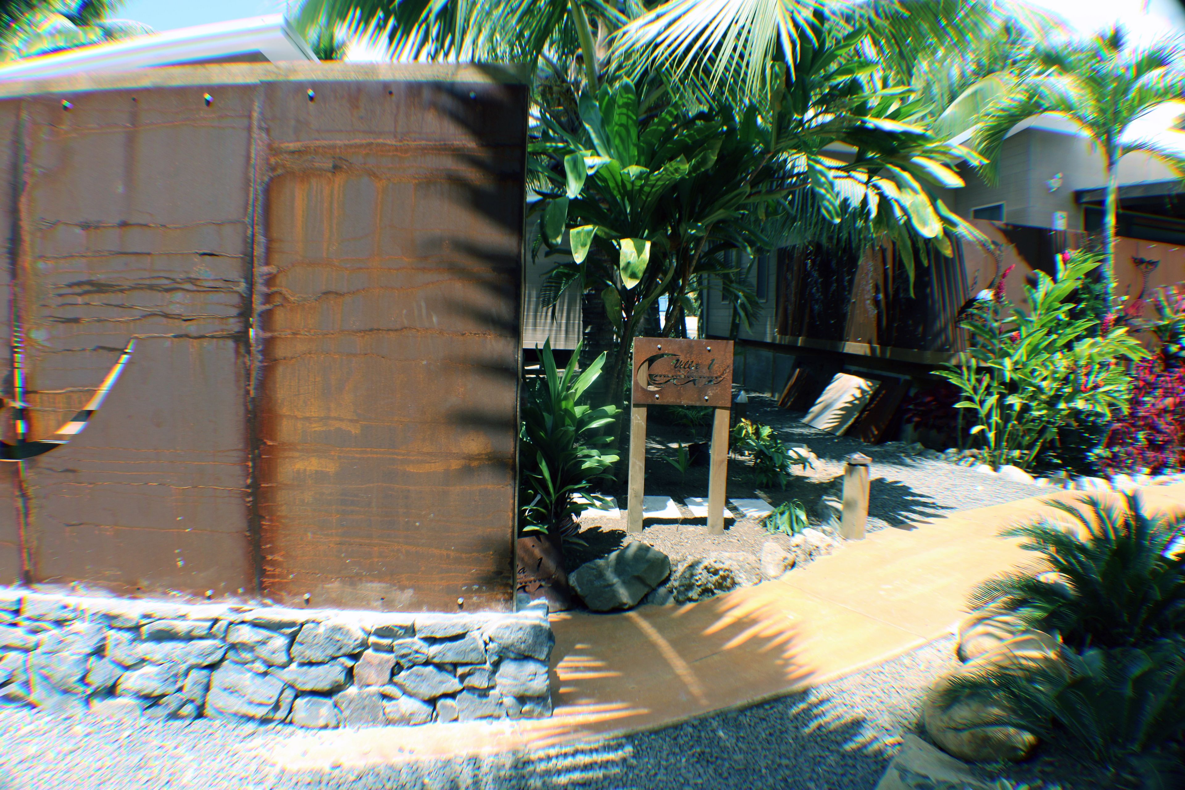 Une structure rustique en bois, dotée d’une base en pierre, entourée de plantes tropicales et agrémentée d’un petit panneau indicateur.