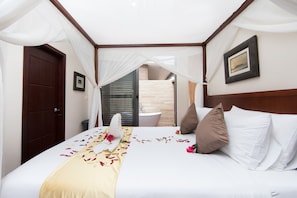 Cama bem arrumada com colcha branca, manta bege e um buquê de flores. Tem uma cabeceira de madeira, uma parede branca e um quadro emoldurado acima da cama.