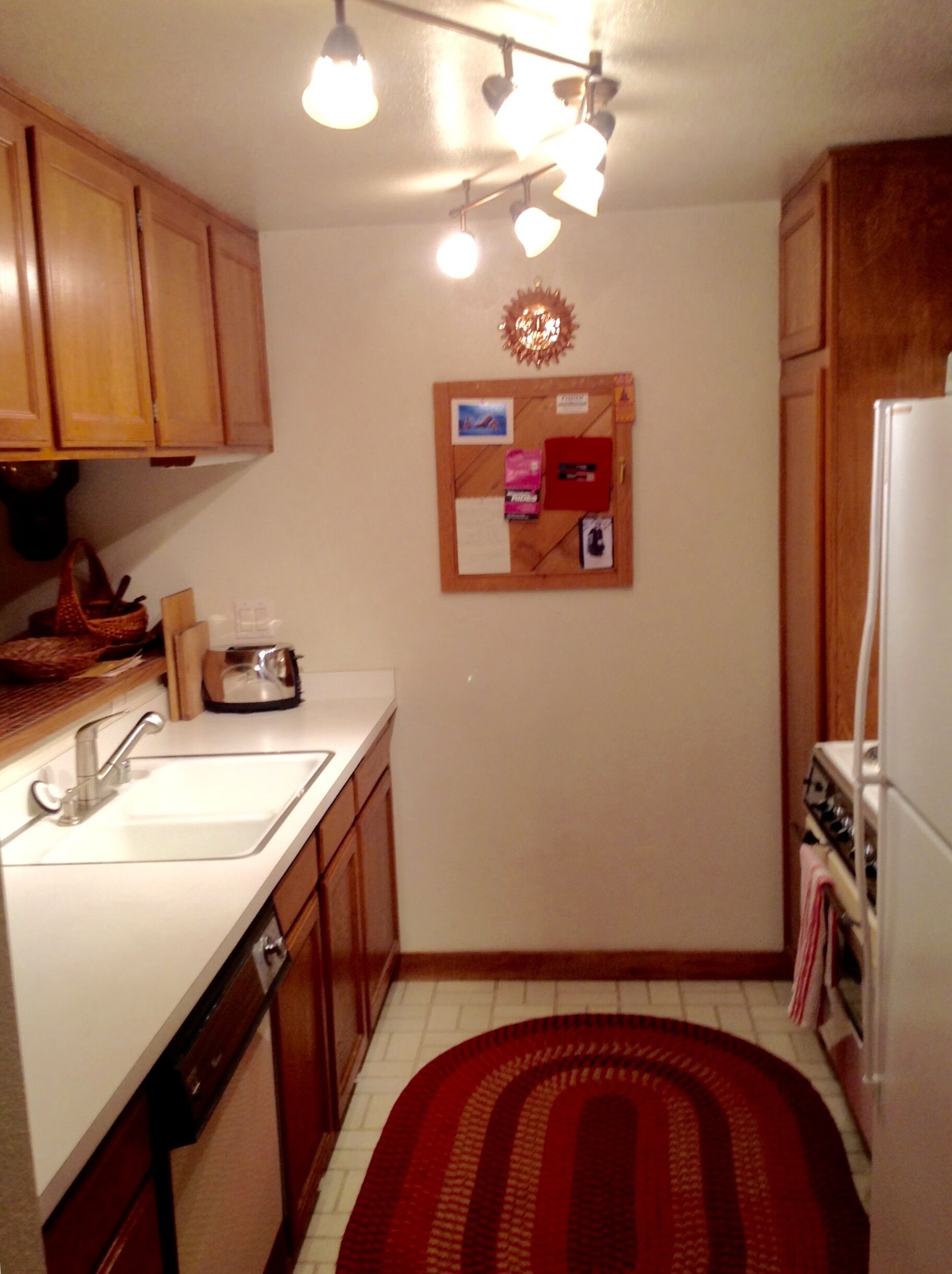 A small kitchen with wooden cabinets, a white sink, a stove, and a corkboard on the wall.