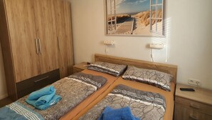 Ein Schlafzimmer mit zwei Betten, einem Holzschrank, einem an der Wand hängenden Bild von einem Strand und einem Nachttisch mit einer Fernbedienung.