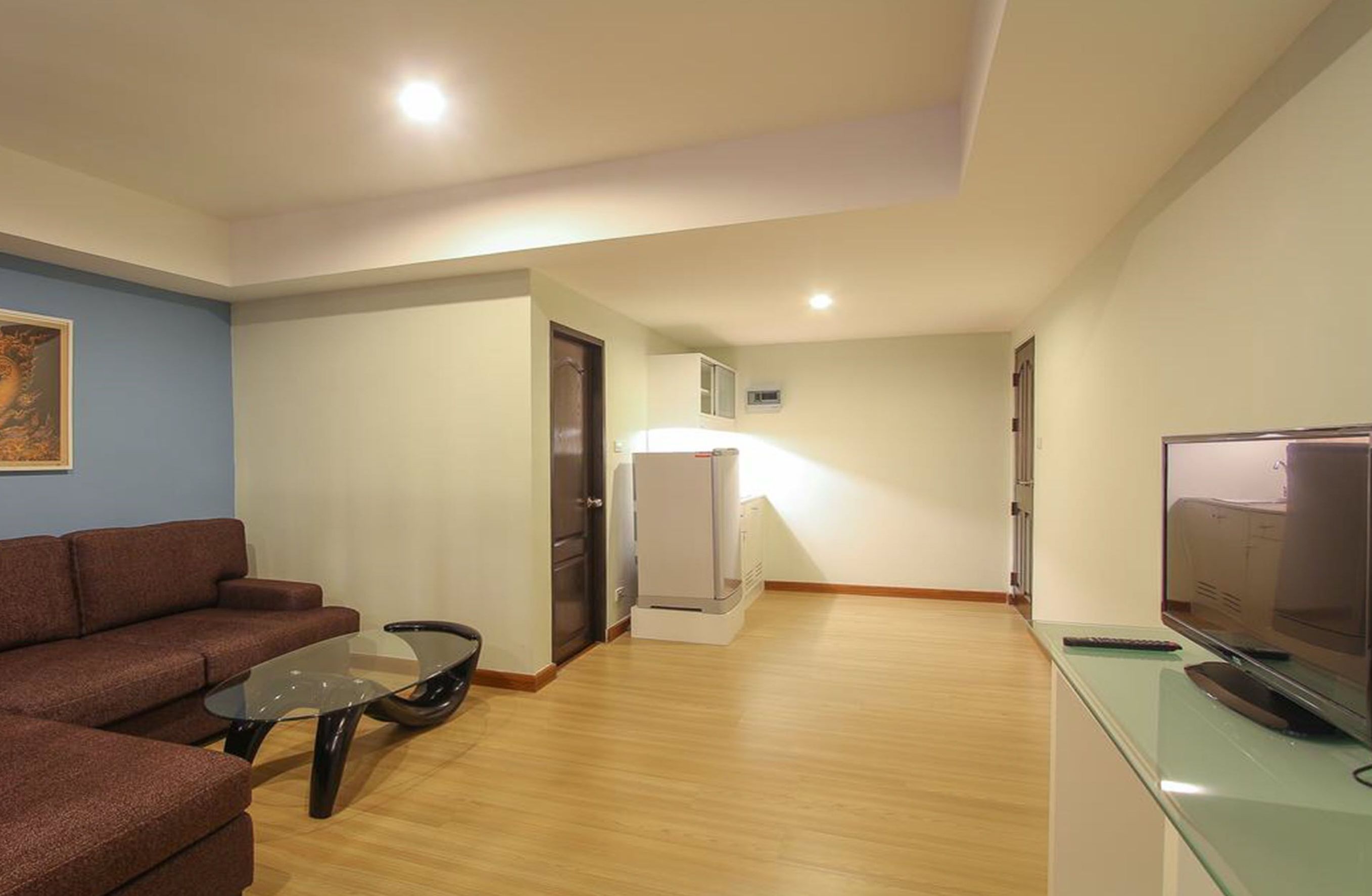 Un salon moderne avec un canapé, une table basse en verre, un téléviseur à écran plat et un réfrigérateur blanc.