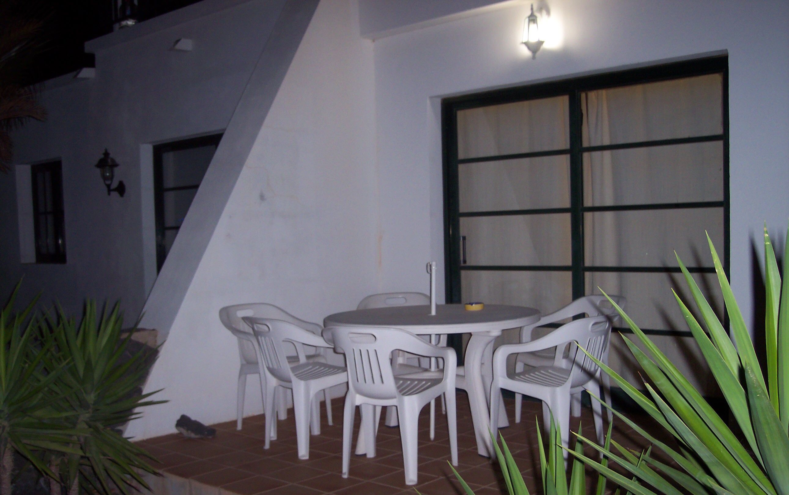 A patio with a round table and four chairs, a window with curtains, and a wall-mounted light fixture.