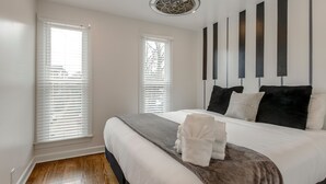 Une chambre moderne avec un grand lit, une literie blanche et un plaid gris. On y trouve deux fenêtres équipées de stores, un ventilateur de plafond et un luminaire fixé au mur.