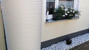 Un grand pot de fleurs blanc, orné de fleurs bleues, placé près d’une fenêtre avec des stores blancs.
