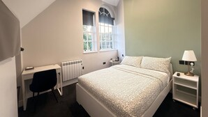 A bedroom with a bed, a desk, a chair, a radiator, a window, and a nightstand with a lamp.