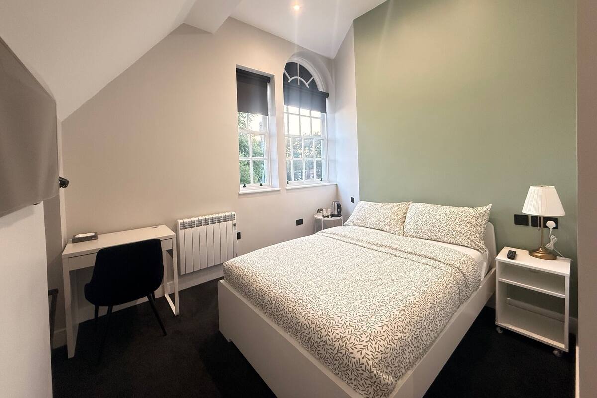 A bedroom with a bed, a desk, a chair, a radiator, a window, and a nightstand with a lamp.